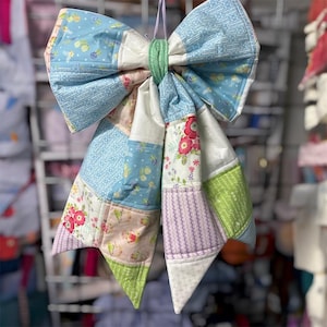 May include: A large, handmade fabric bow with a patchwork design. The bow features various pastel-colored fabrics, including light blue, pink, and green, with floral and patterned prints. A green fabric band secures the center.