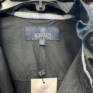 May include: A black jacket featuring a Buffalo David Bitton brand label at the inner neckline with a size M tag. A black string attaches a rectangular beige SewBettySue tag to the garment. The jacket has a ruffled collar and grey trim.