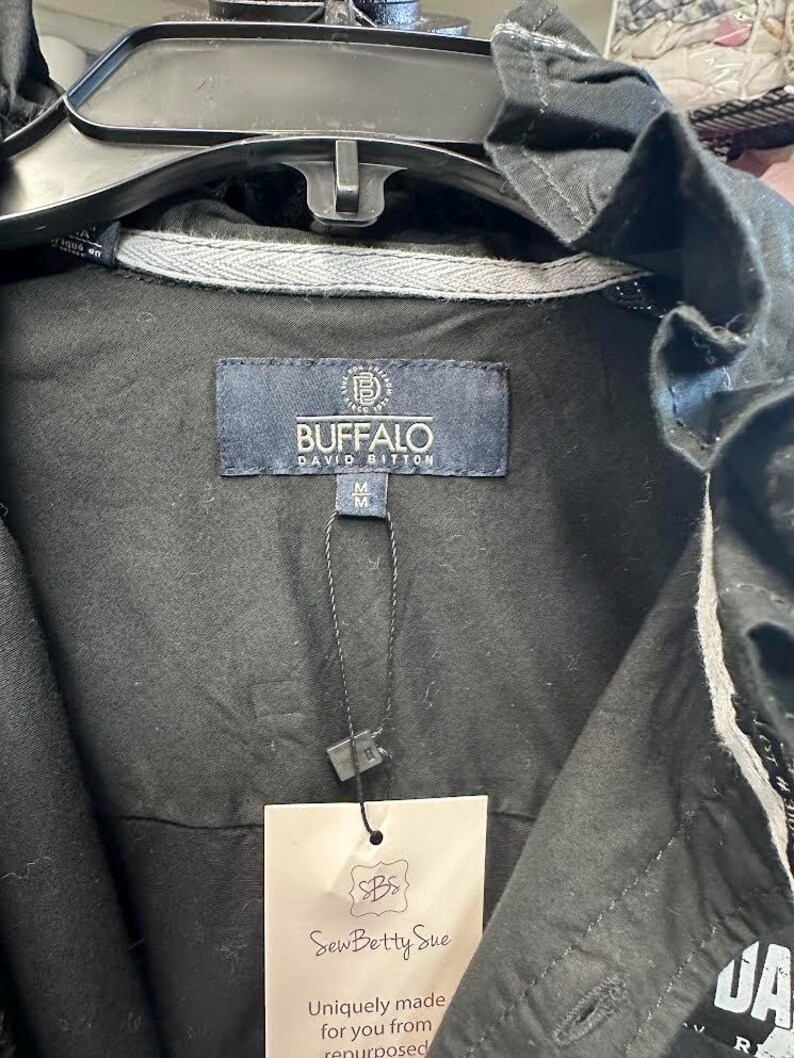May include: A black jacket featuring a Buffalo David Bitton brand label at the inner neckline with a size M tag. A black string attaches a rectangular beige SewBettySue tag to the garment. The jacket has a ruffled collar and grey trim.