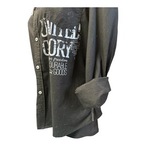 May include: A black long-sleeve button-down shirt featuring white distressed text that reads UNITED CORY for freedom DURABLE GOODS. The shirt has a front pocket, a collar, and sleeves that are rolled up to the elbow, showcasing a casual design.