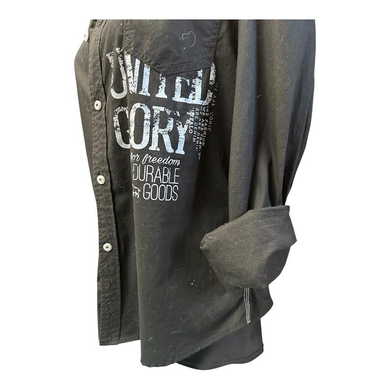 May include: A black long-sleeve button-down shirt featuring white distressed text that reads UNITED CORY for freedom DURABLE GOODS. The shirt has a front pocket, a collar, and sleeves that are rolled up to the elbow, showcasing a casual design.