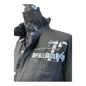 May include: A black button-down shirt featuring a ruffled collar and a front chest pocket. The fabric displays white distressed-style screen-printed text that reads "SINCE 72", "symbol of achievement", and "BUFFALO DAVID BITTON" across the chest area.