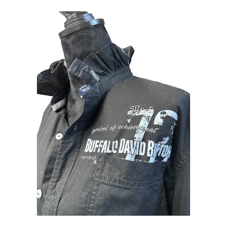 May include: A black button-down shirt featuring a ruffled collar and a front chest pocket. The fabric displays white distressed-style screen-printed text that reads "SINCE 72", "symbol of achievement", and "BUFFALO DAVID BITTON" across the chest area.