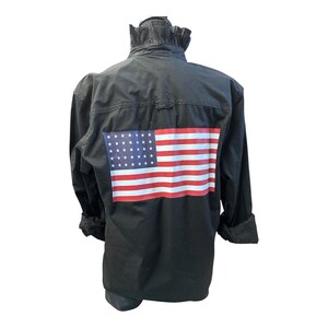 May include: A black long-sleeve shirt featuring a ruffled collar and a rectangular graphic of the American flag printed on the back. The flag design displays horizontal red and white stripes with a blue field containing white stars.