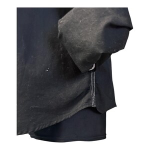 May include: A black long-sleeved shirt featuring a curved hemline and visible white contrast stitching along the side seam. The garment is constructed from a solid black fabric and includes a sleeve with a gathered cuff detail at the wrist area.