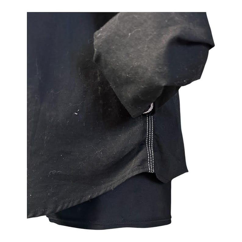 May include: A black long-sleeved shirt featuring a curved hemline and visible white contrast stitching along the side seam. The garment is constructed from a solid black fabric and includes a sleeve with a gathered cuff detail at the wrist area.