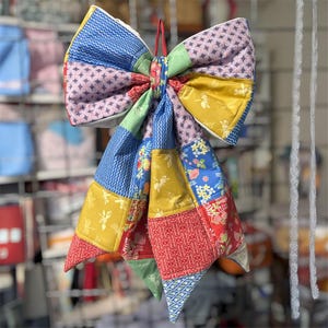 May include: A large, colorful fabric bow made from patchwork squares. The bow features a variety of patterns and colors, including blue, yellow, red, and pink. A red string is used to hang the bow.