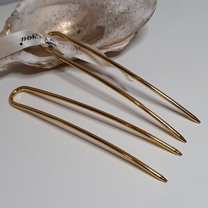Op de afbeelding: Drie gouden haarspelden, elk met een afgerond uiteinde en een puntige punt. De spelden zijn in een verspringende volgorde op een wit oppervlak gerangschikt.