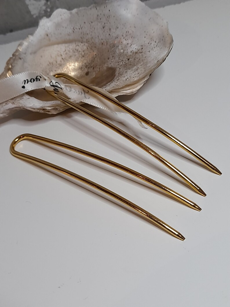 Op de afbeelding: Drie gouden haarspelden, elk met een afgerond uiteinde en een puntige punt. De spelden zijn in een verspringende volgorde op een wit oppervlak gerangschikt.