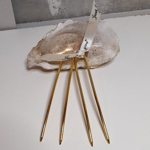 Op de afbeelding: Een witte oesterschelp met een gouden binnenkant, versierd met twee witte satijnen linten met de tekst "Just you" geschreven in zwarte inkt. Vier gouden metalen haarspelden zijn in de schelp geplaatst.