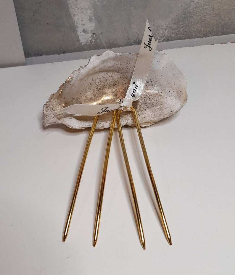 Op de afbeelding: Een witte oesterschelp met een gouden binnenkant, versierd met twee witte satijnen linten met de tekst "Just you" geschreven in zwarte inkt. Vier gouden metalen haarspelden zijn in de schelp geplaatst.