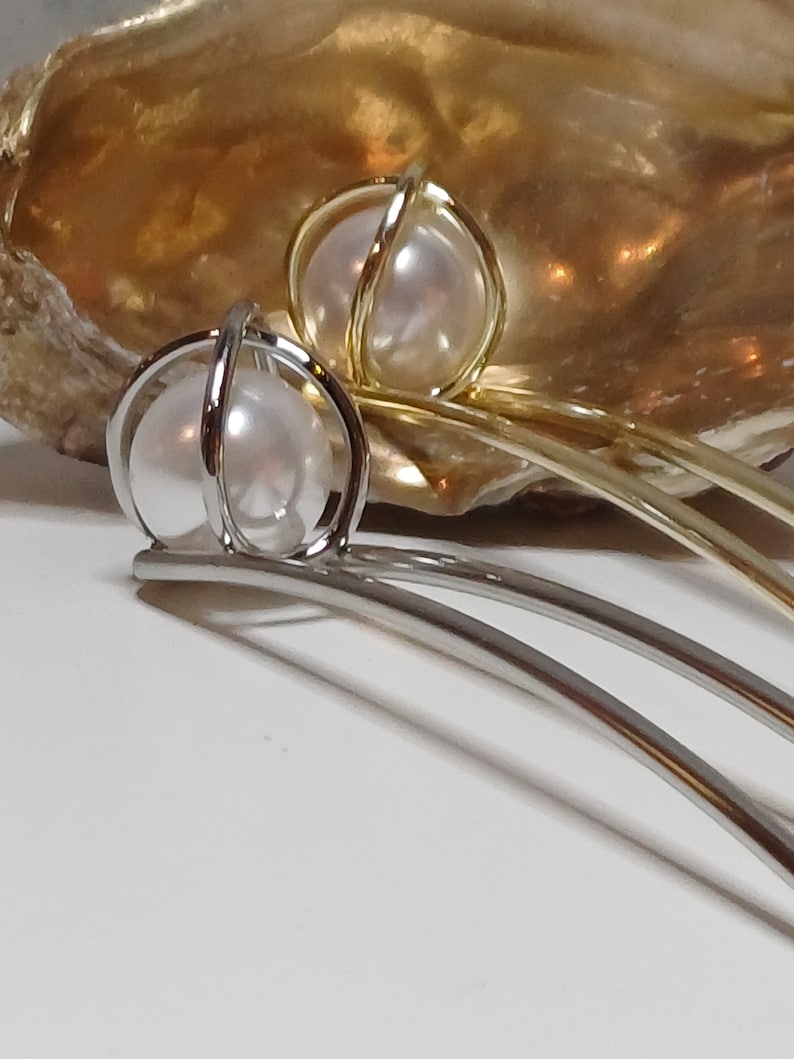 Puede incluir: Dos pinzas para el pelo plateadas con una perla encerrada en una jaula de alambre. Las pinzas est&aacute;n sobre una superficie blanca.