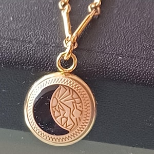 Könnte beinhalten: Eine goldfarbene Halskette mit einem kreisförmigen Anhänger. Der Anhänger zeigt ein Halbmond-Design mit schwarzem Hintergrund und einem Sonnenmuster. Die Kette besteht aus kleinen, verbundenen Gliedern.