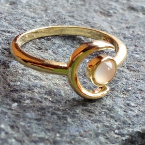 May include: A gold ring with a crescent moon design and a white gemstone. The ring is on a gray textured surface.
