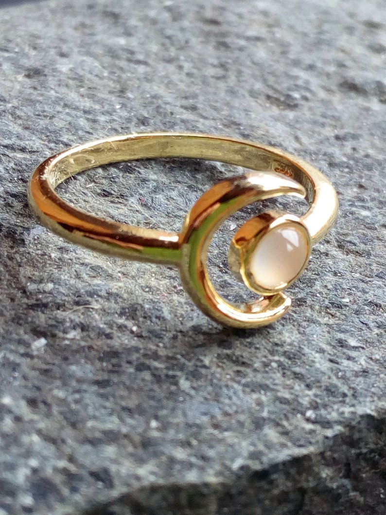 May include: A gold ring with a crescent moon design and a white gemstone. The ring is on a gray textured surface.