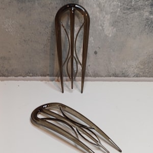 Könnte beinhalten: Zwei dunkelbraune Haarkämme. Einer steht aufrecht und zeigt eine gebogene Oberseite mit dekorativen Linien und drei Zinken. Der andere liegt, mit einem ähnlichen Design. Die Kämme bestehen aus einem durchscheinenden Material.