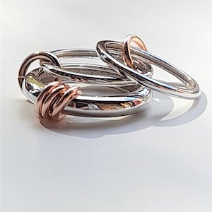 Könnte beinhalten: Drei ineinandergreifende Ringe, ein silberner und zwei roségoldene, liegen auf einer weißen Oberfläche. Die Ringe sind schlicht und elegant.