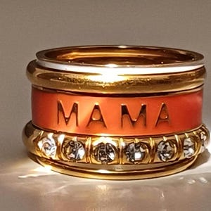 Könnte beinhalten: Ein goldener Ring mit einem roten Band, auf dem "MAMA" steht, und einer Reihe von klaren Kristallen.