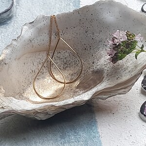Könnte beinhalten: Ein Paar goldene Ohrringe in Tropfenform mit einer zarten Kette, die von der Oberseite jedes Ohrrings hängt. Die Ohrringe werden in einer weißen Muschelschale präsentiert.