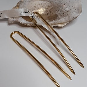 Op de afbeelding: Drie gouden haarspelden, ook wel haarsticks genoemd, liggen op een wit oppervlak. De spelden zijn dun en hebben een lichte bocht.