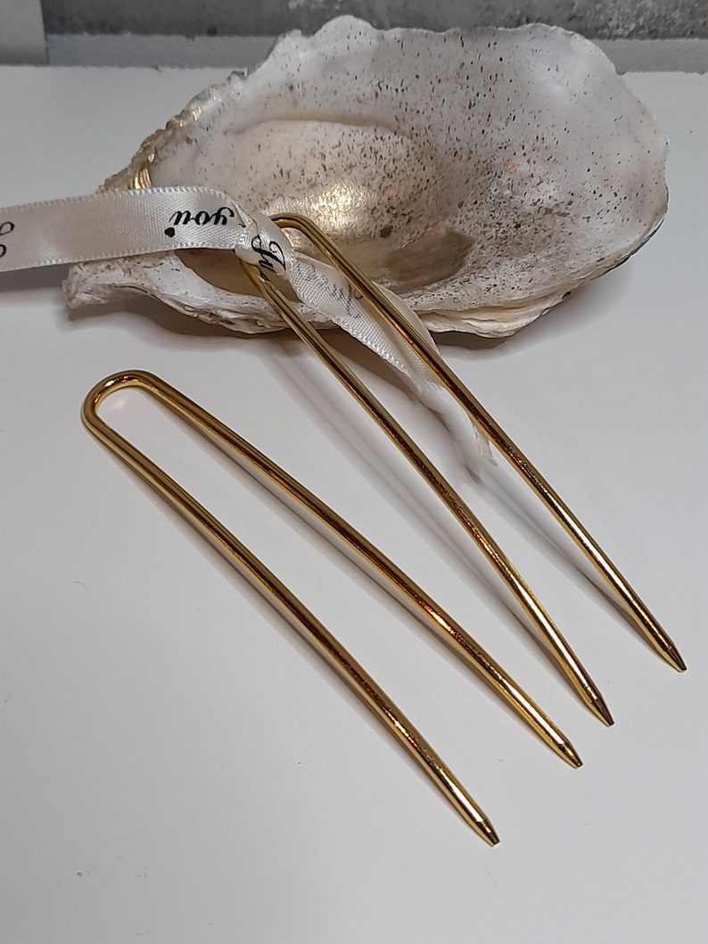 Op de afbeelding: Drie gouden haarspelden, ook wel haarsticks genoemd, liggen op een wit oppervlak. De spelden zijn dun en hebben een lichte bocht.