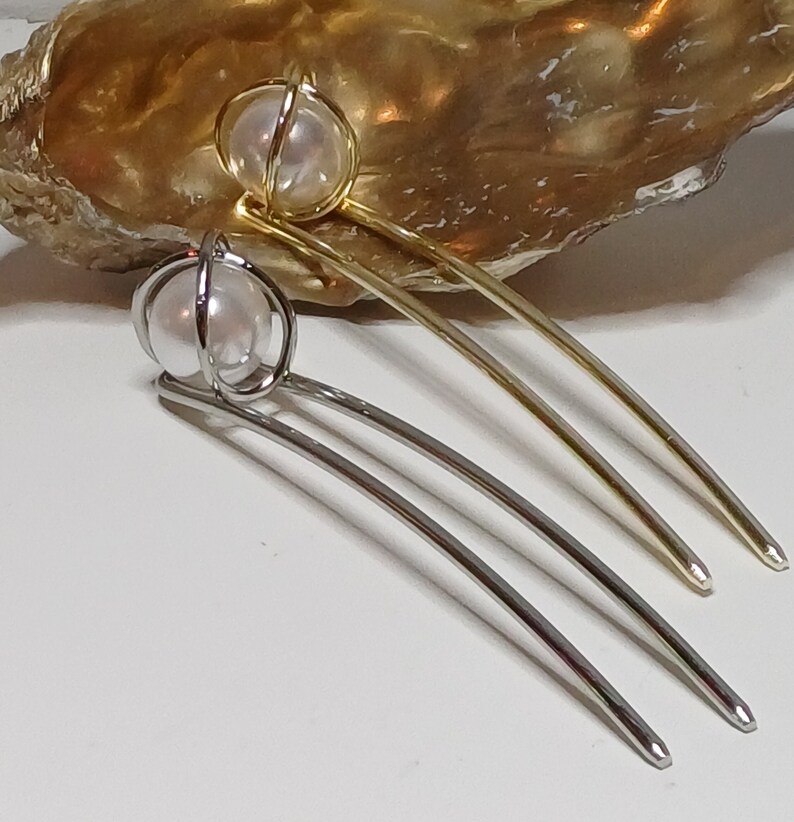 Puede incluir: Dos pinzas para el pelo con detalles de perlas. Las pinzas son de metal y tienen una forma curva. Una pinza es dorada y la otra es plateada.