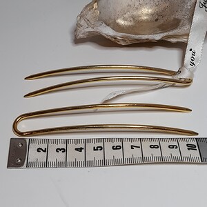 Op de afbeelding: Drie gouden haarspelden, twee recht en een met een gebogen uiteinde, liggen op een wit oppervlak naast een meetlint. Het lint meet 25,4 cm.