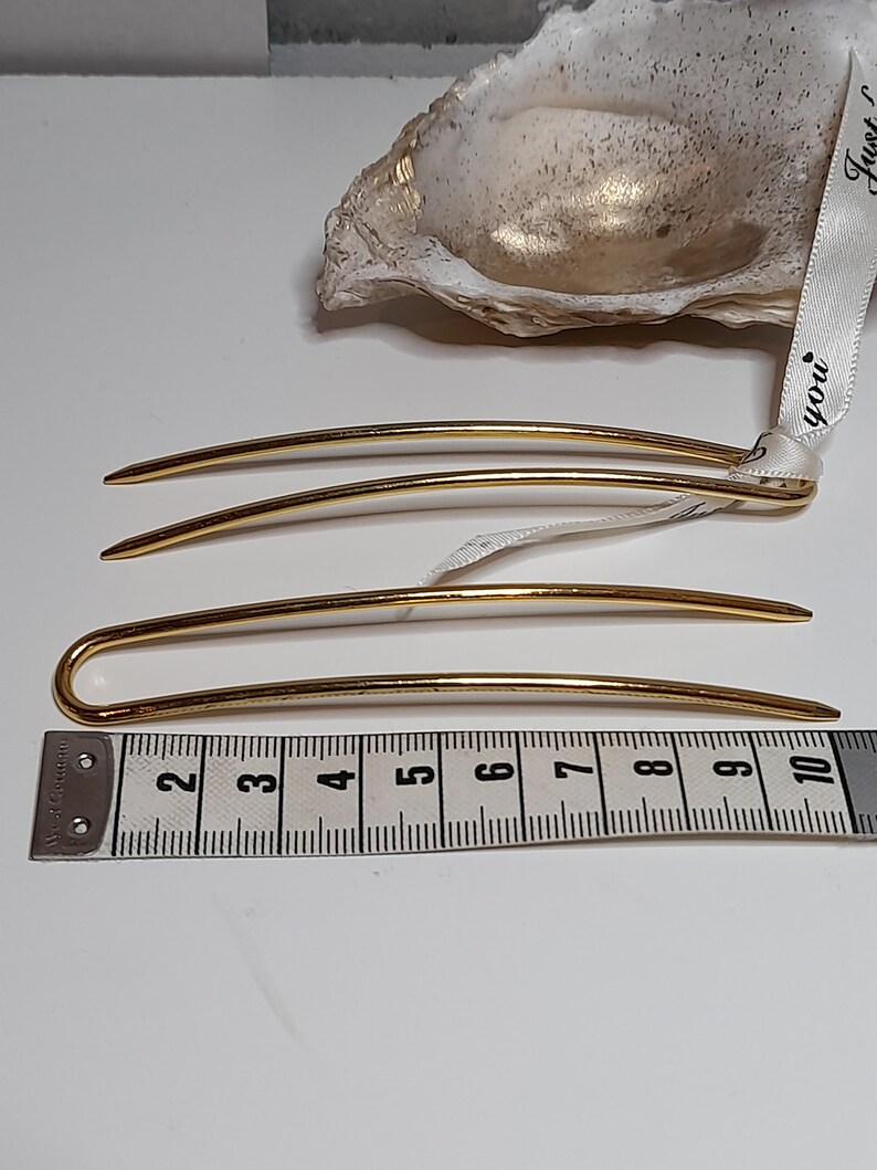 Op de afbeelding: Drie gouden haarspelden, twee recht en een met een gebogen uiteinde, liggen op een wit oppervlak naast een meetlint. Het lint meet 25,4 cm.