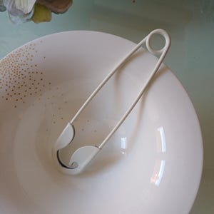Op de afbeelding: Een grote, gebroken witte veiligheidsspeld met een gladde, matte afwerking. De speld is voorzien van een klassiek spiraalveer-mechanisme aan het ene uiteinde en een veilige sluiting aan het andere, ontworpen voor het vastzetten van stof of als accessoire.