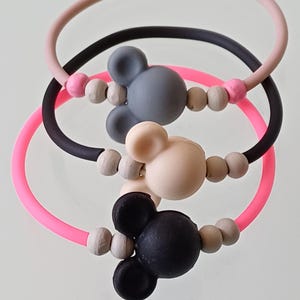 May include: Three colorful silicone bracelets with Mickey Mouse-shaped beads. The bracelets are pink, black, and light pink, each with a different colored mouse head. The beads are a mix of light gray and pink.