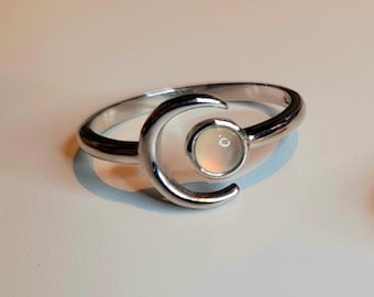 May include: A silver-toned ring featuring a crescent moon design that curves around a small, round, translucent white stone set in the center. The metal band is smooth and polished, creating a reflective surface that catches the light.