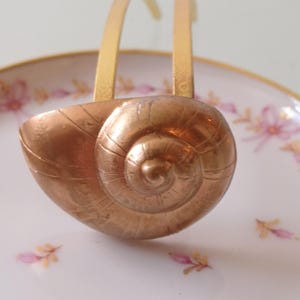 Könnte beinhalten: Eine dekorative Servierschale mit einem kupferfarbenen, schneckenförmigen Griff. Die Schale hat eine weiße Oberfläche mit einem floralen Muster in Rosa und Gelb. Der Griff ist aus goldfarbenem Metall.