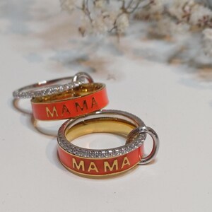Könnte beinhalten: Zwei goldene Ringe mit einem roten Emailleband und einer Reihe von klaren Steinen. Die Ringe sagen "MAMA" in goldenen Buchstaben.