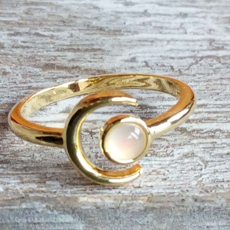May include: A gold crescent moon ring with a white opal gemstone. The ring is adjustable and can be worn on any finger.