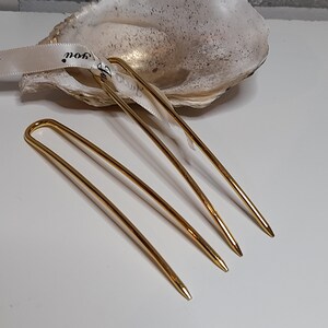 Op de afbeelding: Drie gouden haarspelden, elk met een licht verschillende bocht, liggen op een wit oppervlak. De spelden zijn ongeveer 12,5 cm lang.