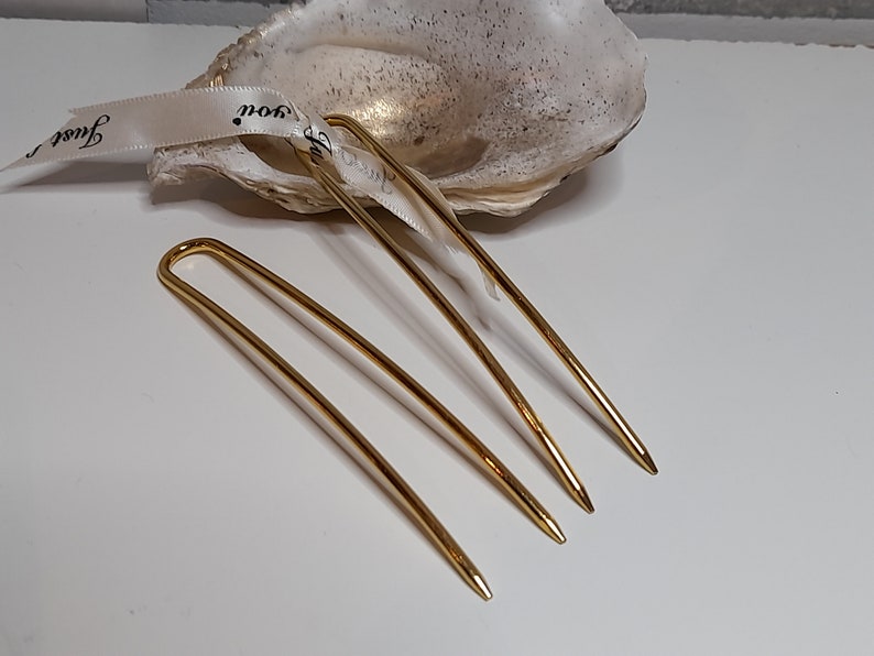 Op de afbeelding: Drie gouden haarspelden, elk met een licht verschillende bocht, liggen op een wit oppervlak. De spelden zijn ongeveer 12,5 cm lang.