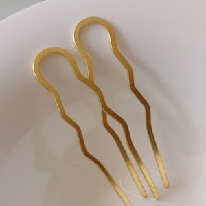 Könnte beinhalten: Zwei goldfarbene Haarnadeln mit gewelltem Design. Die Nadeln bestehen aus glänzendem Metall und haben eine U-förmige Oberseite mit drei Zinken unten. Die Nadeln liegen auf einer weißen Oberfläche.