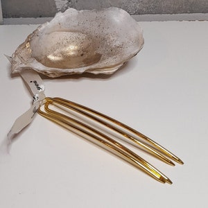 Op de afbeelding: Twee gouden haarspelden met een lus aan de bovenkant. De spelden liggen op een wit oppervlak.