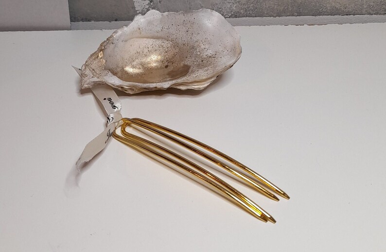 Op de afbeelding: Twee gouden haarspelden met een lus aan de bovenkant. De spelden liggen op een wit oppervlak.