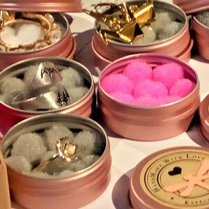 May include: Assortment of small pink metal tins with various items inside. The tins have a gold rim and are filled with white, pink, and gold items. One tin has a ring with a pink stone, another has a ring with a silver pyramid, and another has pink pom-poms.