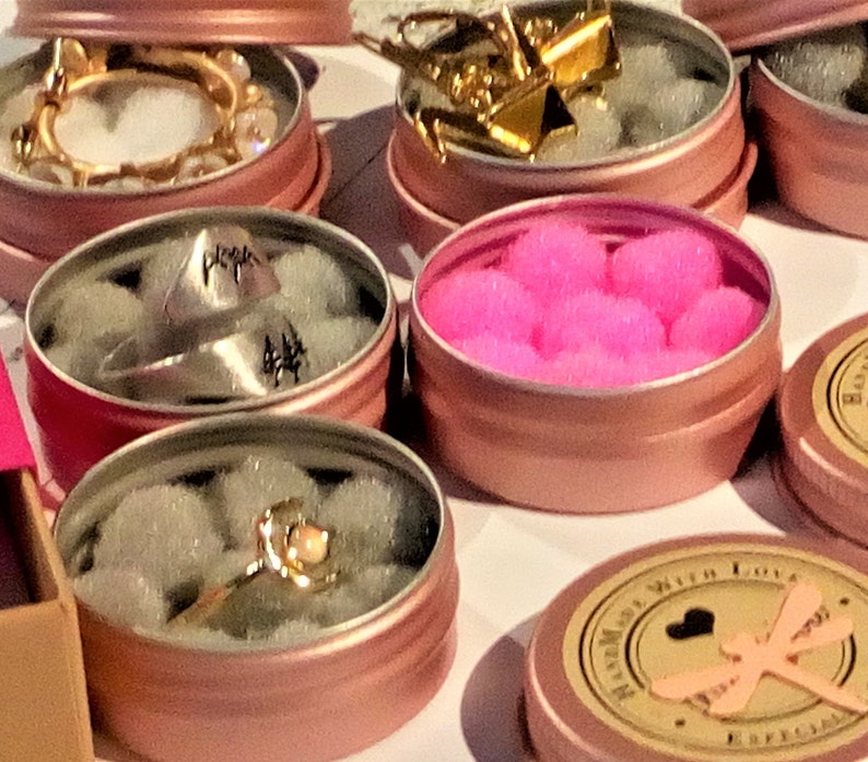 May include: Assortment of small pink metal tins with various items inside. The tins have a gold rim and are filled with white, pink, and gold items. One tin has a ring with a pink stone, another has a ring with a silver pyramid, and another has pink pom-poms.