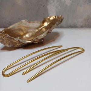 Op de afbeelding: Drie gouden haarspelden, elk met een licht verschillende bocht, liggen op een wit oppervlak. De spelden zijn gemaakt van glad, glanzend metaal.