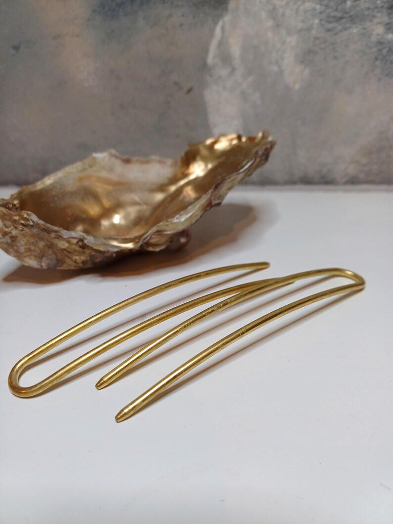 Op de afbeelding: Drie gouden haarspelden, elk met een licht verschillende bocht, liggen op een wit oppervlak. De spelden zijn gemaakt van glad, glanzend metaal.