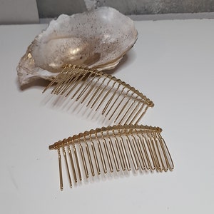 Könnte beinhalten: Zwei goldene Haarkämme mit einer geschwungenen Form und einer Reihe von Zähnen. Die Kämme liegen auf einer weißen Oberfläche.