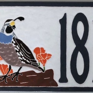 May include: A white ceramic house number sign with the number 18105 in black. The sign features a painted image of a quail with blue and brown feathers and orange flowers on either side.