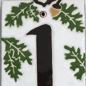 Puede incluir: Un azulejo de cerámica blanco con números negros 17940 y hojas de roble verdes con bellotas marrones. El azulejo es un número de casa.