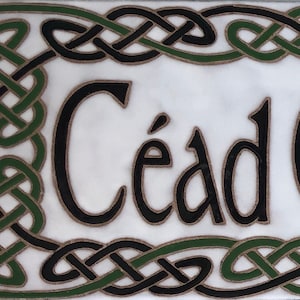 Könnte beinhalten: Ein weißes Keramik-Schild mit einem schwarzen und grünen keltischen Knotenrand. Das Schild trägt die Aufschrift "Cead Mile Failte" in schwarzen Buchstaben.