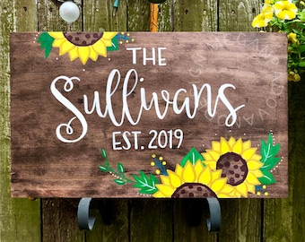 May include: A wooden sign with the text "The Sullivans Est. 2019" painted in white. The sign is decorated with painted sunflowers and green leaves.