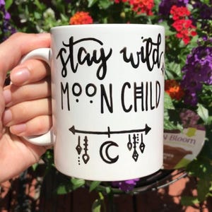 A person's hand holding a white mug with the text "Stay wild moon child" printed on it, against a background of colorful flowers.