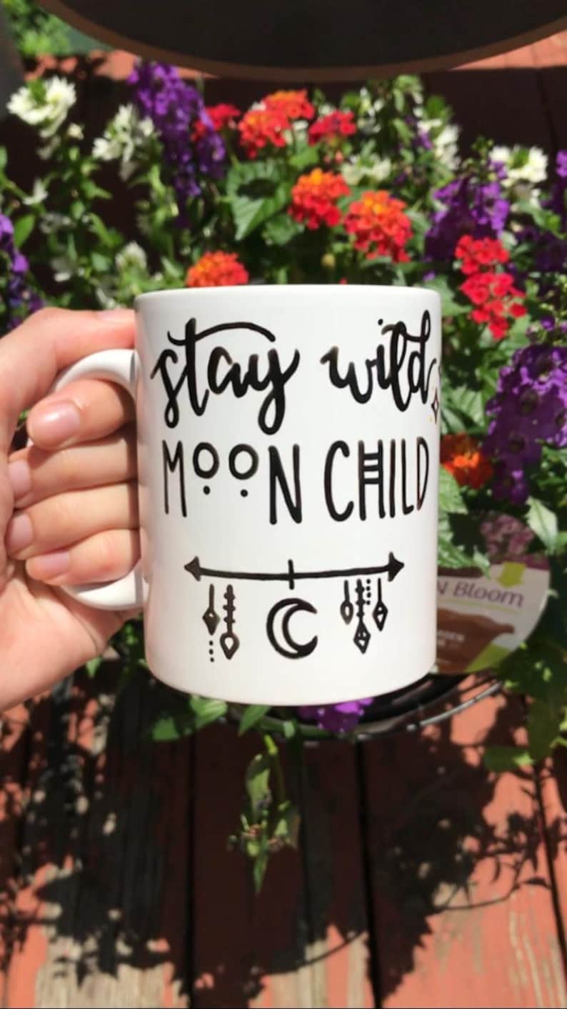 A person's hand holding a white mug with the text "Stay wild moon child" printed on it, against a background of colorful flowers.